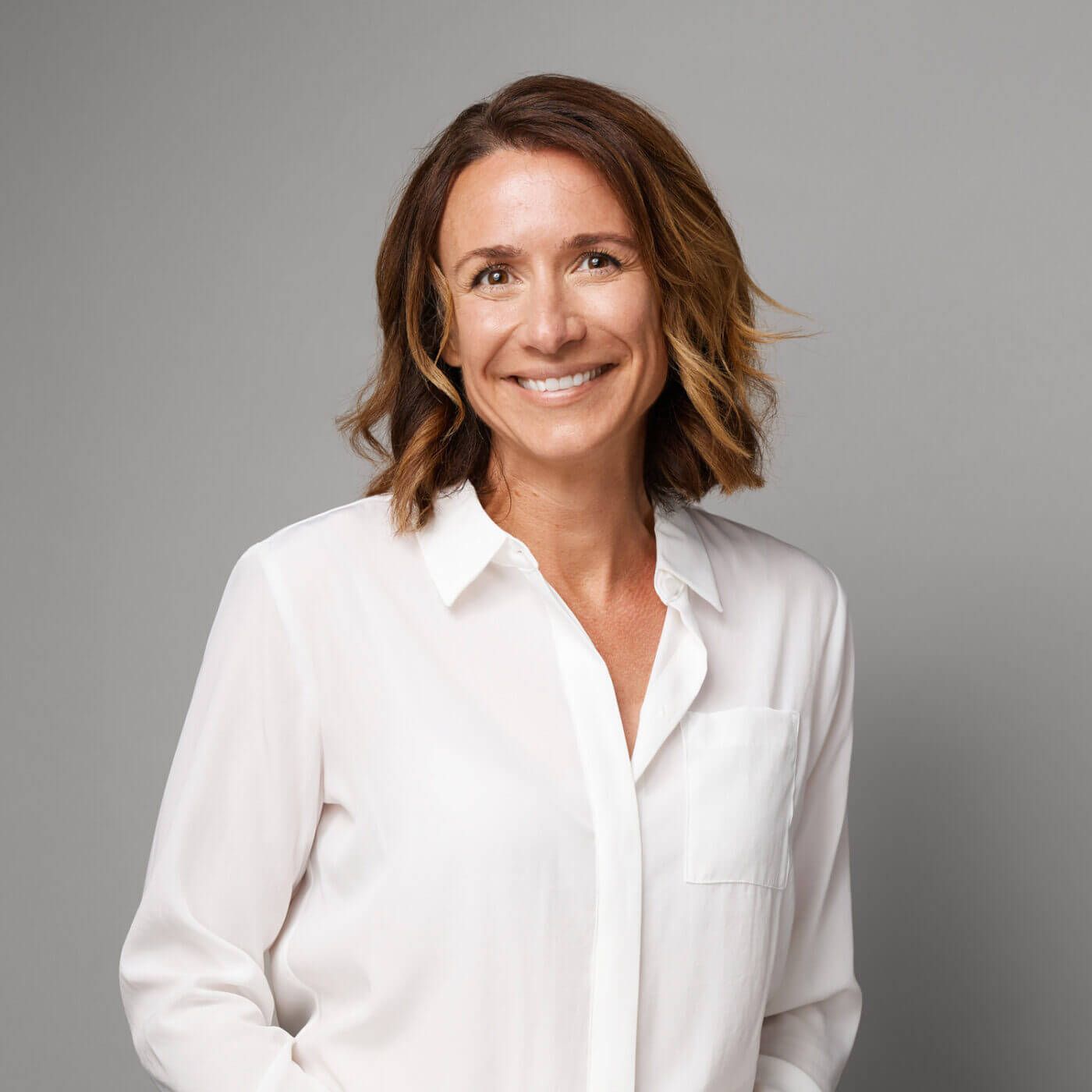 A white woman with shoulder-length brown hair wears a white collared shirt and smiles at the camera
