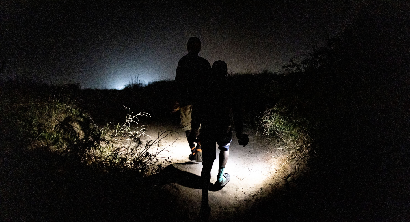 Two shadowed people shine a bright flashlight on the ground as they walk through a dark night. Dawn breaks on the horizon line