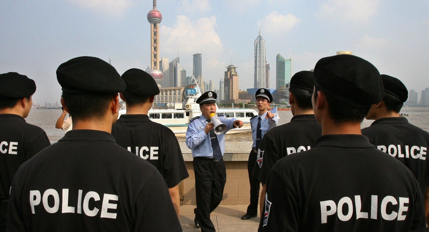 Several police stand with their backs to the camera wearing plain black shirts with the word "POLICE" printed in white block letters. Two police in blue uniforms address the crowd. They stand in front of a city skyline.