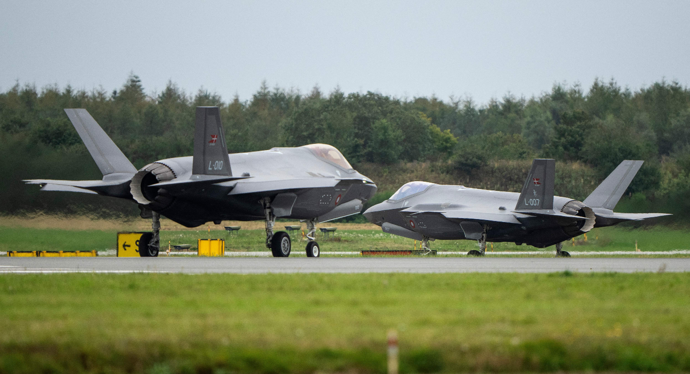 Two fighter jet planes parked on a runway. Lush green grass in foreground and large green trees in background