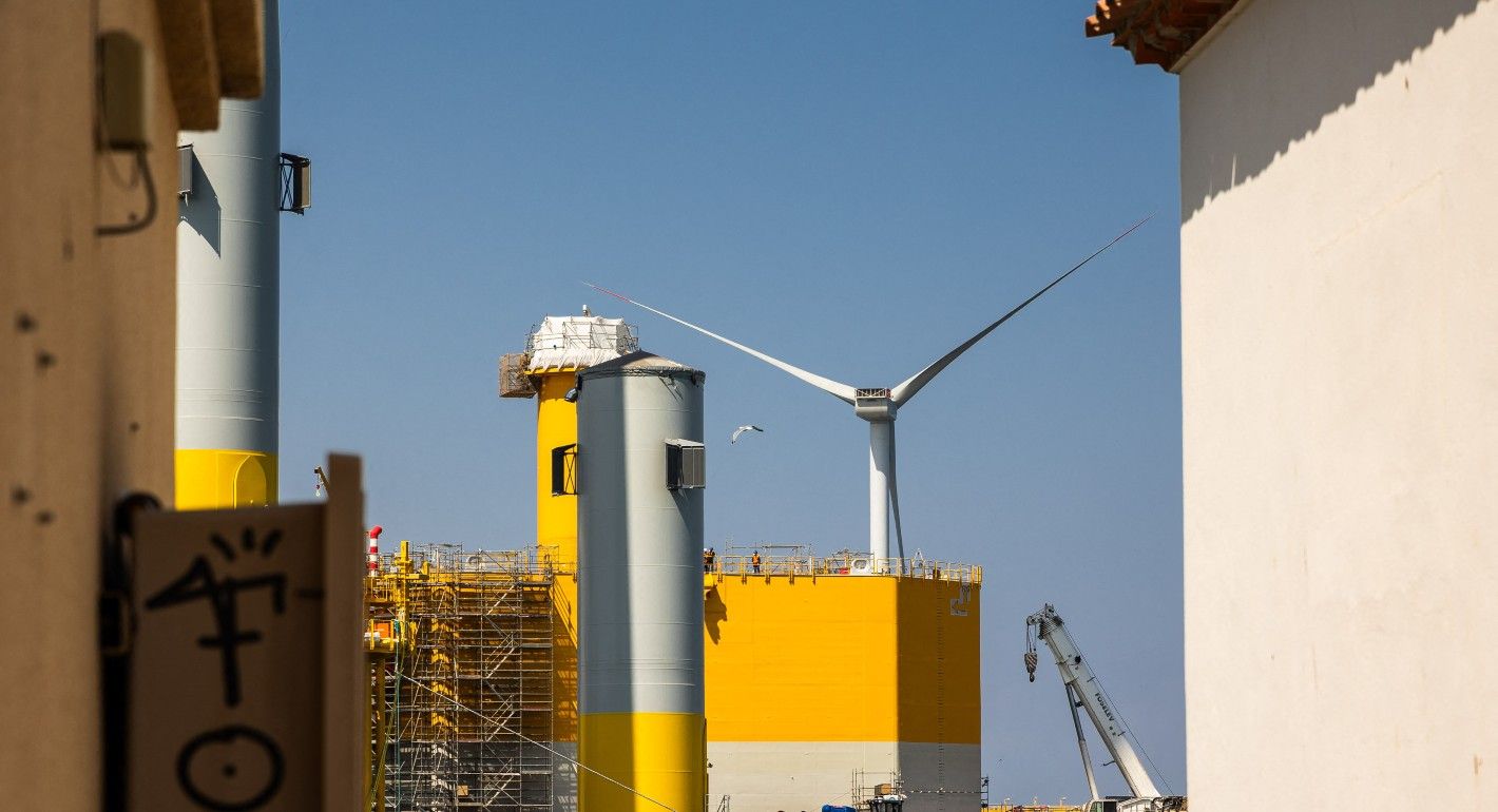 A wind turbine stands behind large yellow-painted manufacturing buildings in front of a blue sky