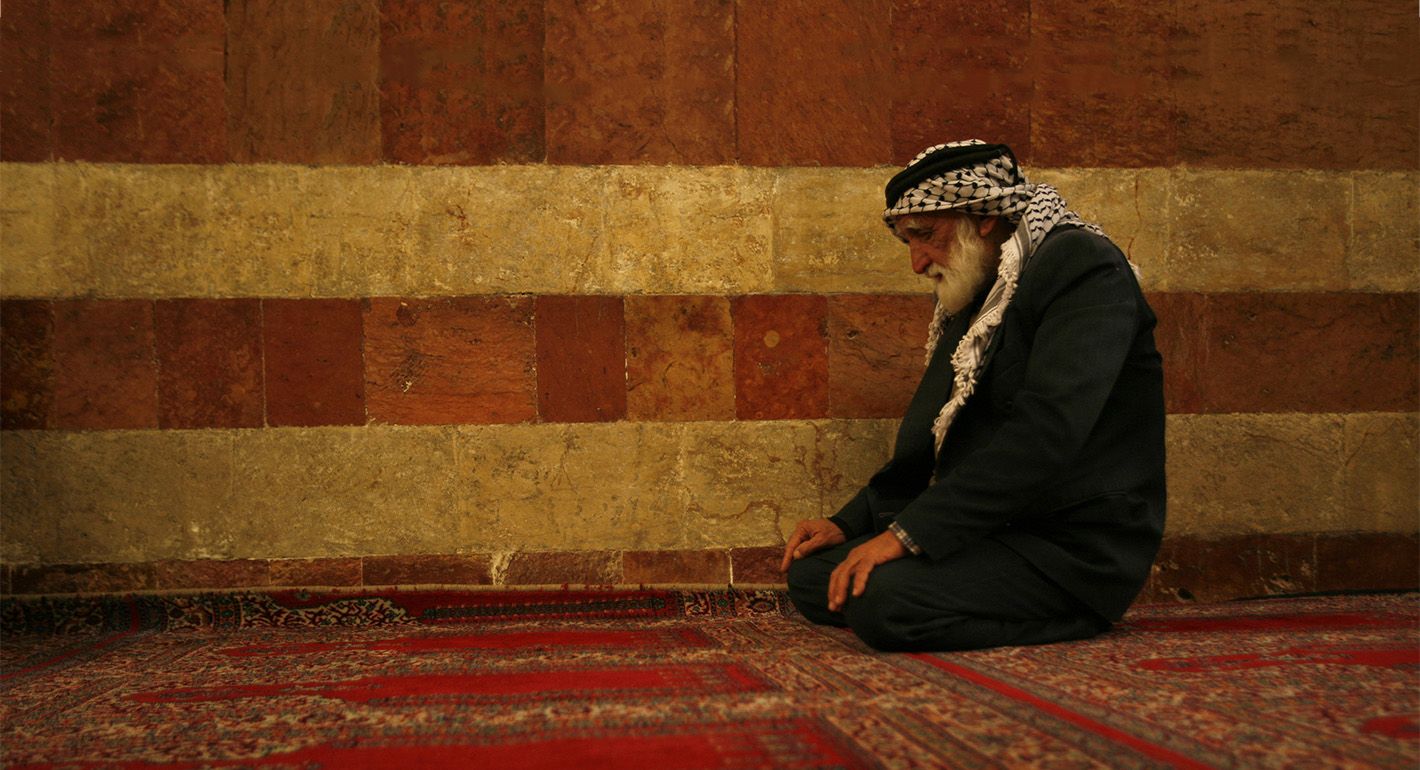 Older man wearing Palestinian keffiyeh kneels on a red and orange prayer rug in front of a red and orange striped mosque wall.