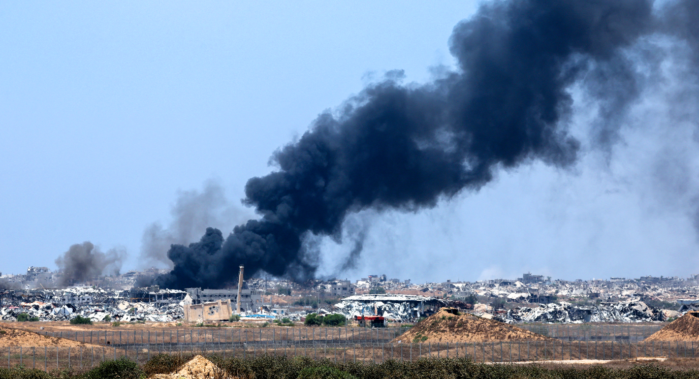 Black plume of smoke stretches across a blue sky from mid left to top right of the photo. Bottom portion shows barren land in foreground and destroyed buildings in the background 