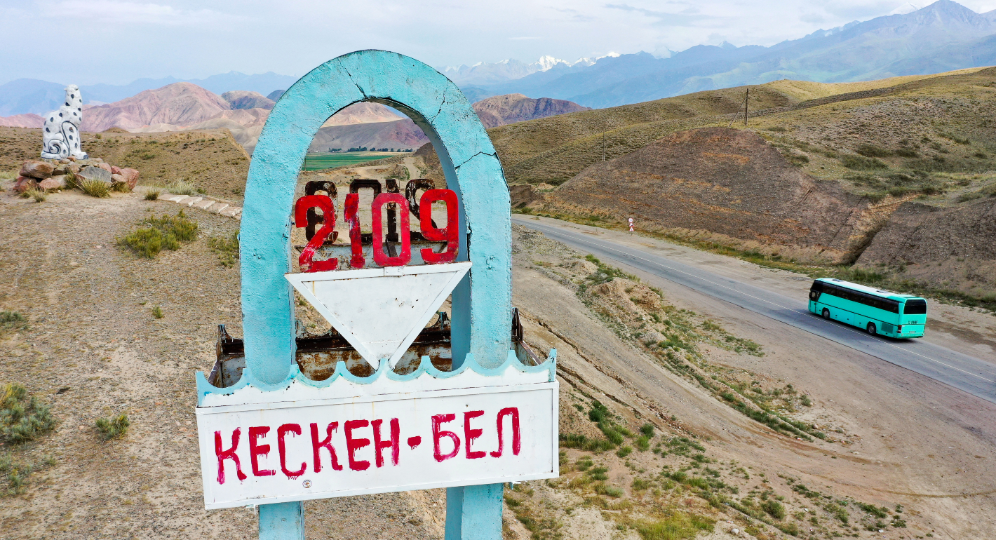 Pale blue arch road sign in foreground in front of a a car riding along the Bishkek - Karakol road along dusty brown mountains