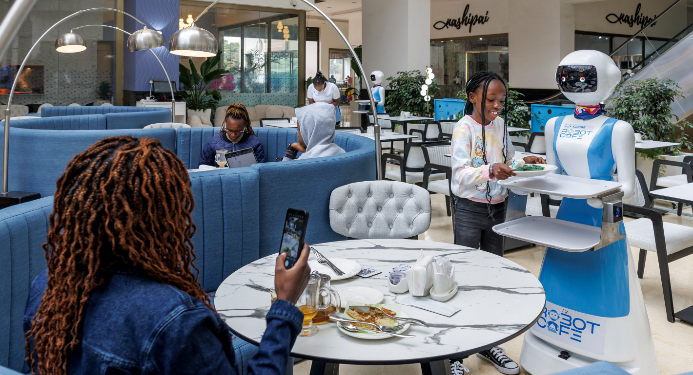 Black woman with auburn braids takes a picture of young Black child smiling standing next to a robot serving lunch at a blue and white restaurant where they are eating