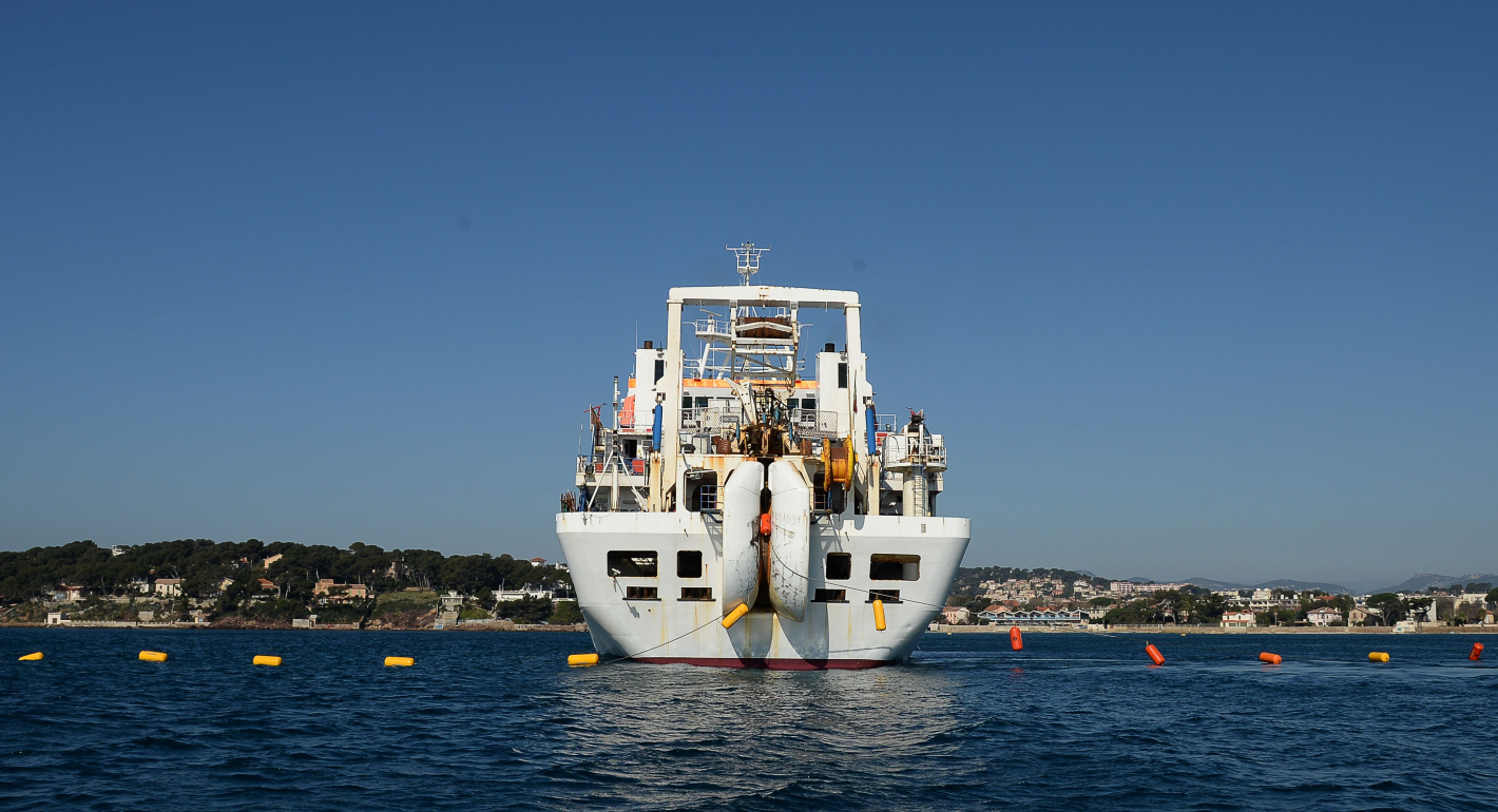 The front of a large white ship is centered on a bright blue sky and blue ocean water. Lush green land and mountains along the horizon line