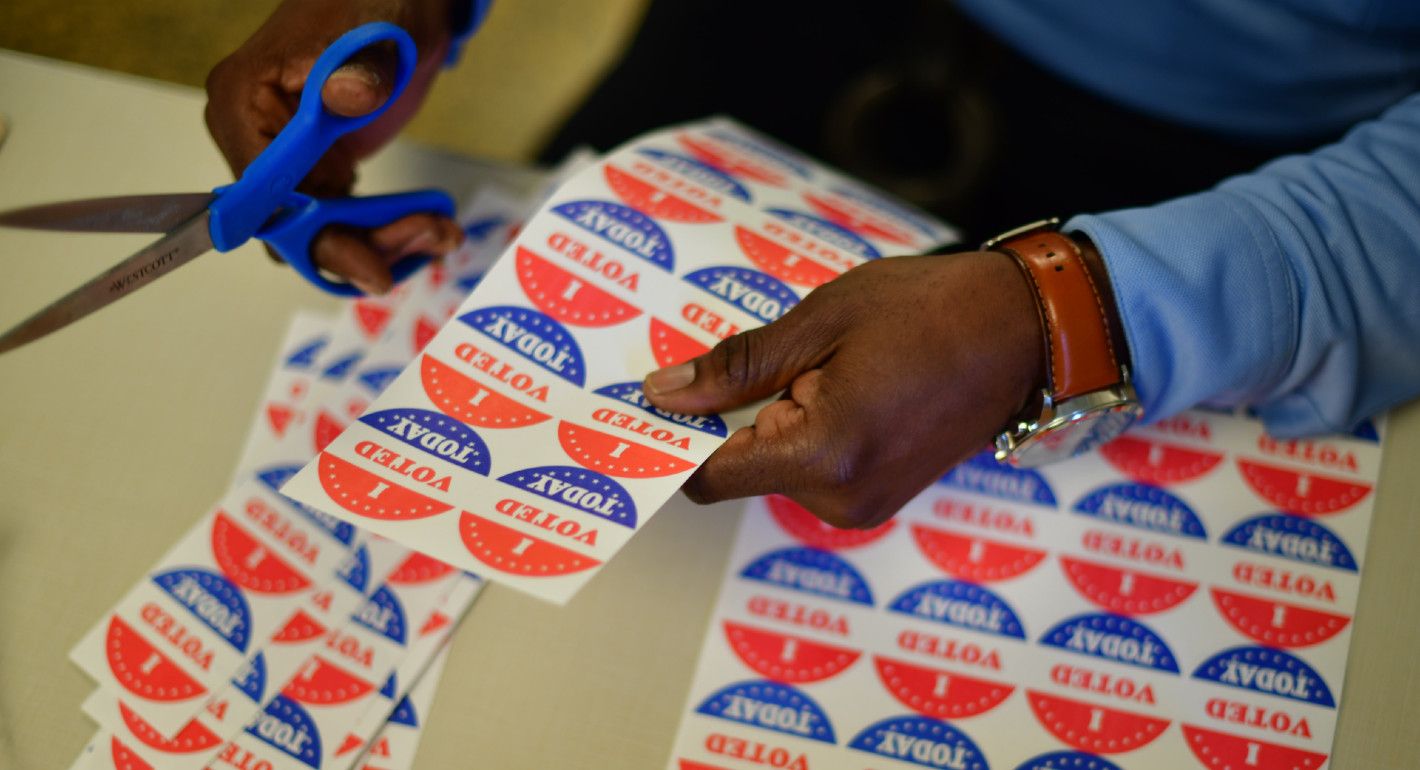 A Black person holds scissors and a sheet of "I Voted" stickers.