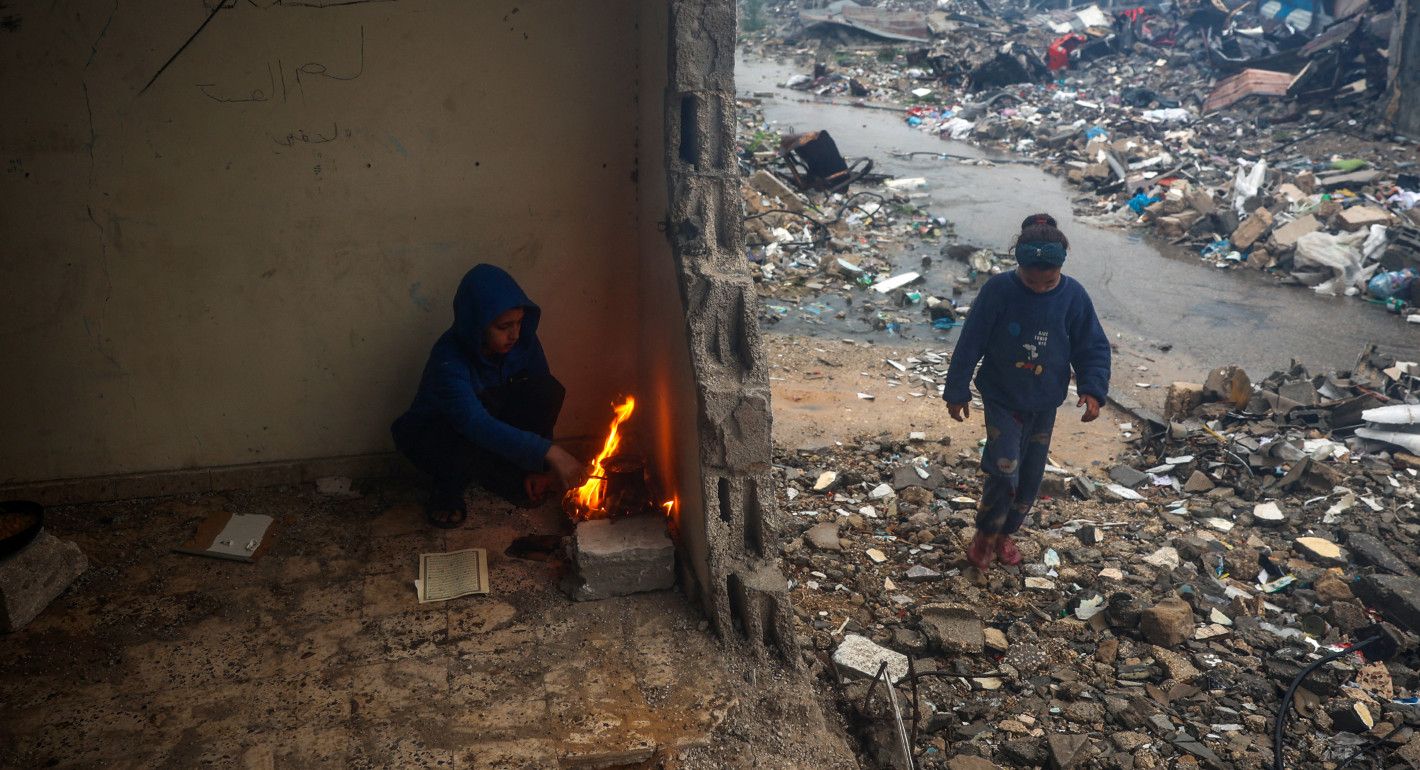 One kid sitting next to a small fire in a damaged building, with another kid walking nearby amid rubble