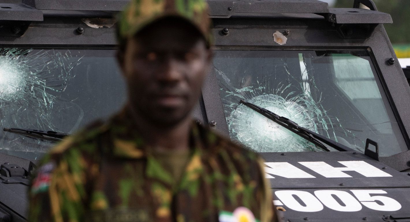 A soldier in the foreground and an armored vehcile with a cracked windshield in the background