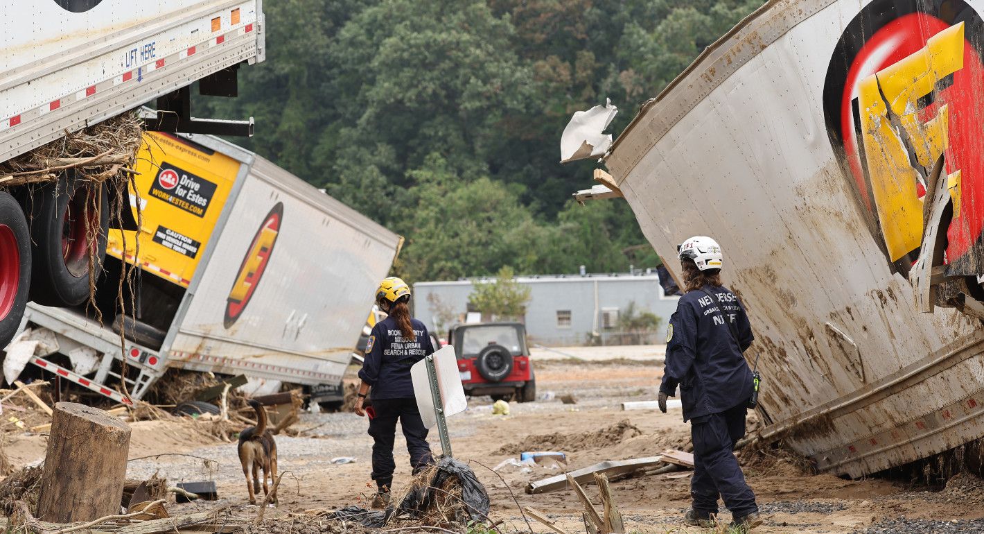 two workers and a dog walking amid ruins of semis damaged in floods