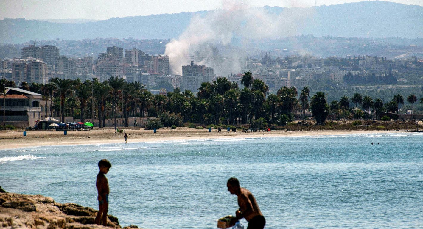 In the foreground, a man and child stand on a beach. In the background, smoke among buildings from an airstrike.