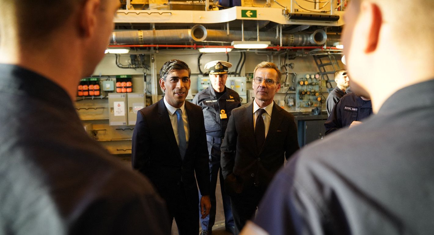 two suited men who are facing the camera and talking to sailors with their backs to the camera on a ship