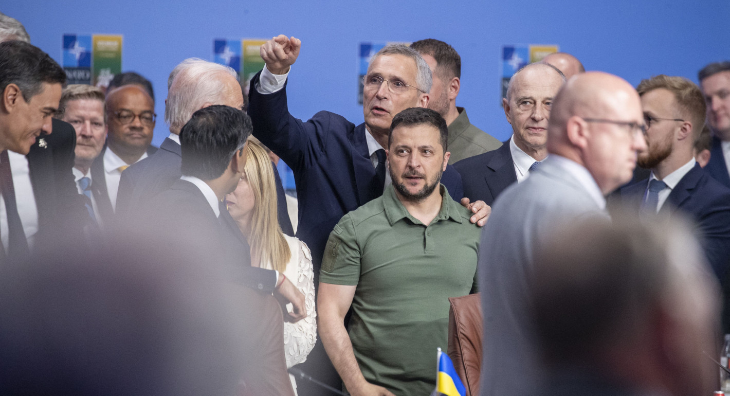Stoltenberg stands over Zelensky, pointing, with both are surrounded by many other people