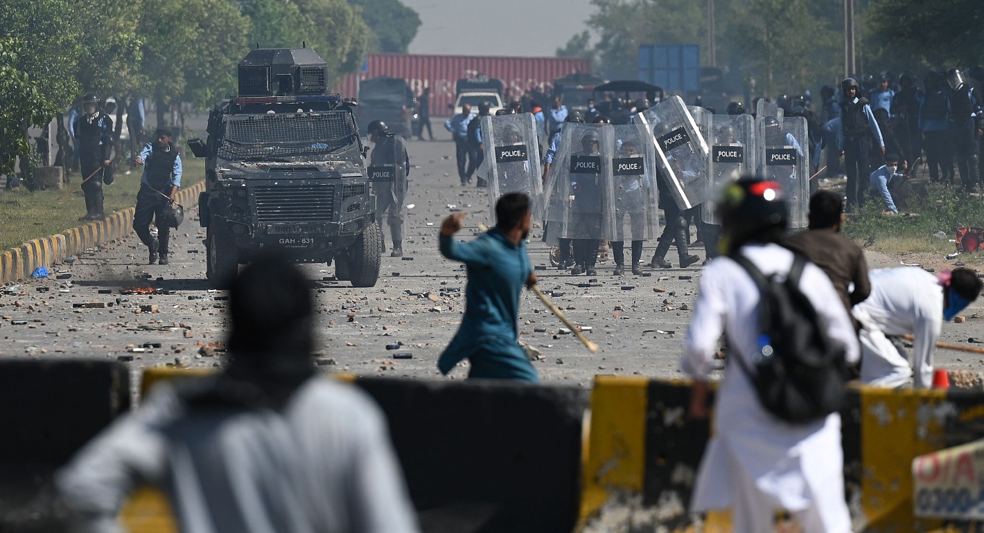 Pakistani police with riot shields advance toward protesters behind a makeshift barricade. One protester stands outside the barricade with arm upraised.