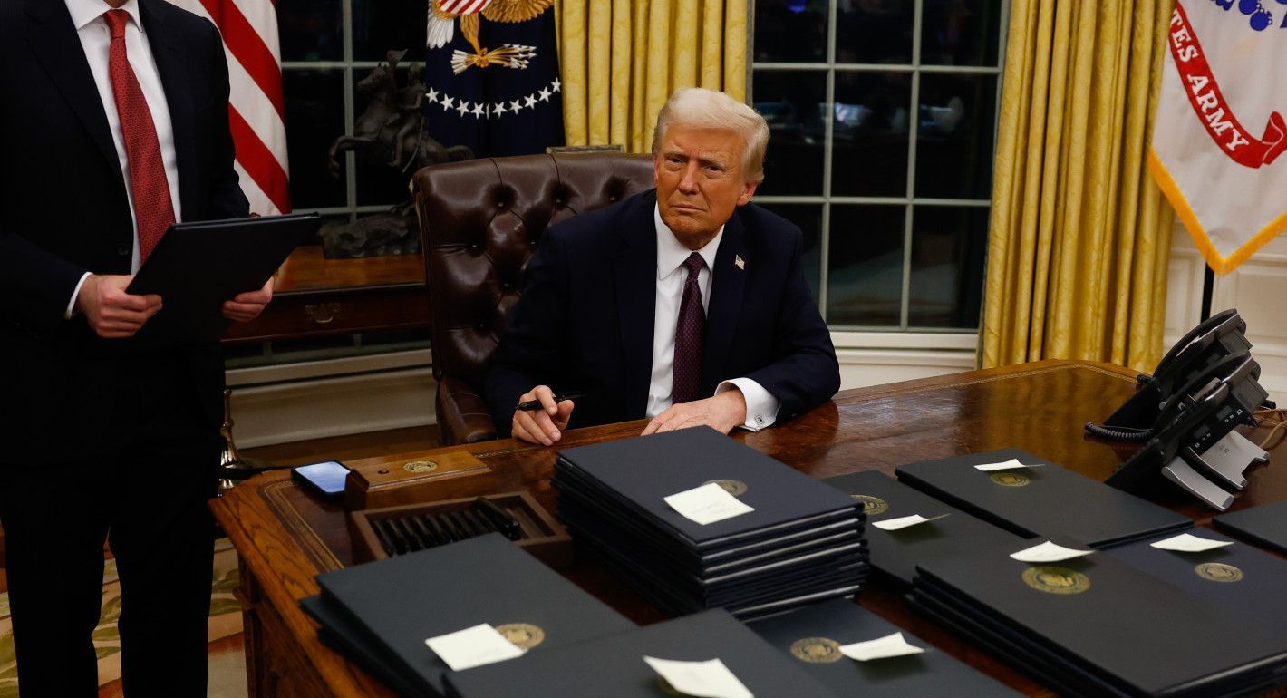 Trump sitting at a desk, pen in hand, with piles in folders in front of him