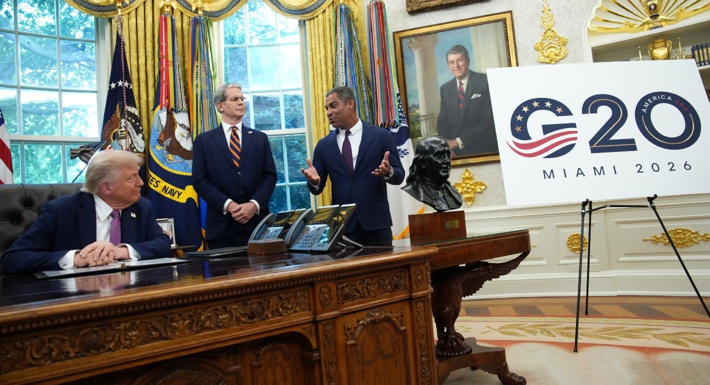 Trump sitting behind a desk while to other men talk